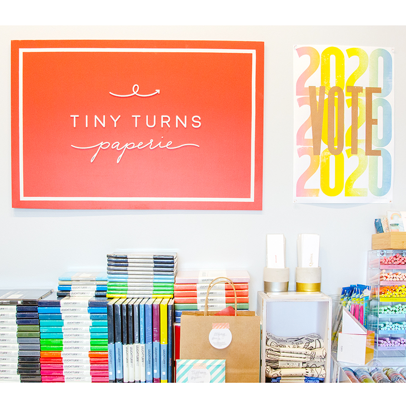 Photo of back wall and counter of Tiny Turns Paperie with notebooks, bookmarks and pens on the counter. The Tiny Turns Paperie logo is prominent on the back wall with a Vote 2020 poster beside it.